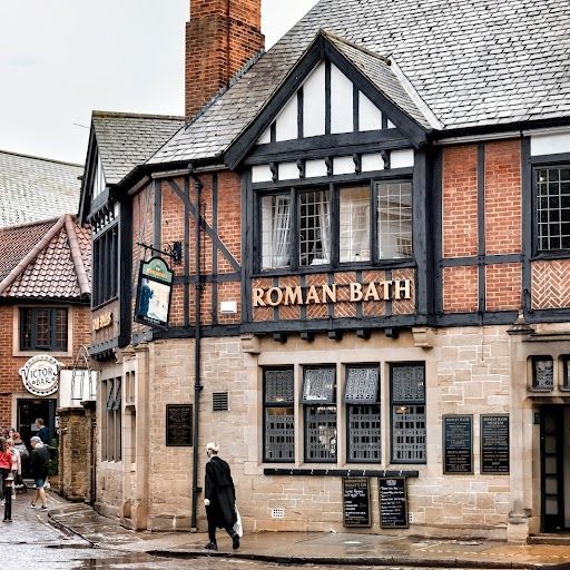 This image captures the exterior of a building, showcasing its architectural details. The composition is well-balanced, drawing the viewer's eye to the prominent "Roman Bath" sign. The lighting is adequate, although slightly overcast, resulting in a soft and even illumination. The image is sharp, revealing the intricate details of the brickwork, windows, and sign. The colors are accurate, portraying the natural hues of the building materials. The image is free from unwanted elements, such as clutter or text. However, the presence of a person walking in the foreground might be considered an unwanted element based on the provided criteria. Overall, the image is a good representation of the building's exterior, showcasing its architectural features and ambiance. The score is slightly reduced due to the presence of the person in the foreground.