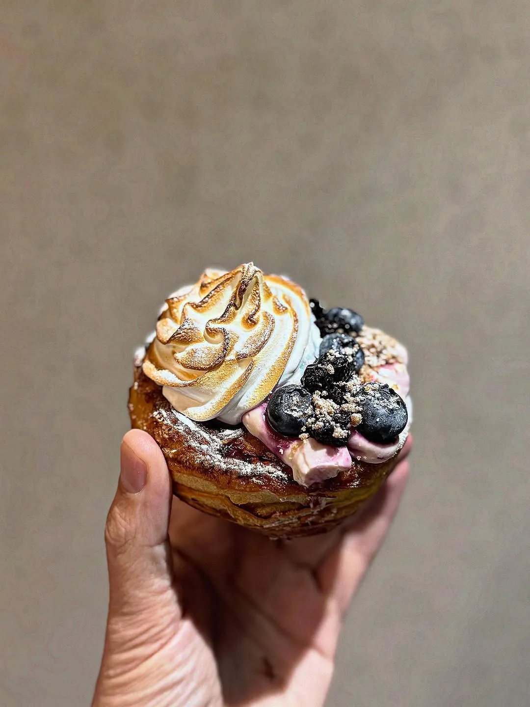 A visually appealing close-up of a pastry topped with toasted meringue and fresh blueberries, held in a hand. The dessert features a golden-brown pastry base, a swirl of perfectly toasted meringue, and a scattering of dark blue blueberries and a light dusting of powder. The background is a soft, neutral tone, ensuring the food is the central focus. The image effectively highlights the textures and colors of the ingredients, making the dessert look highly appetizing.