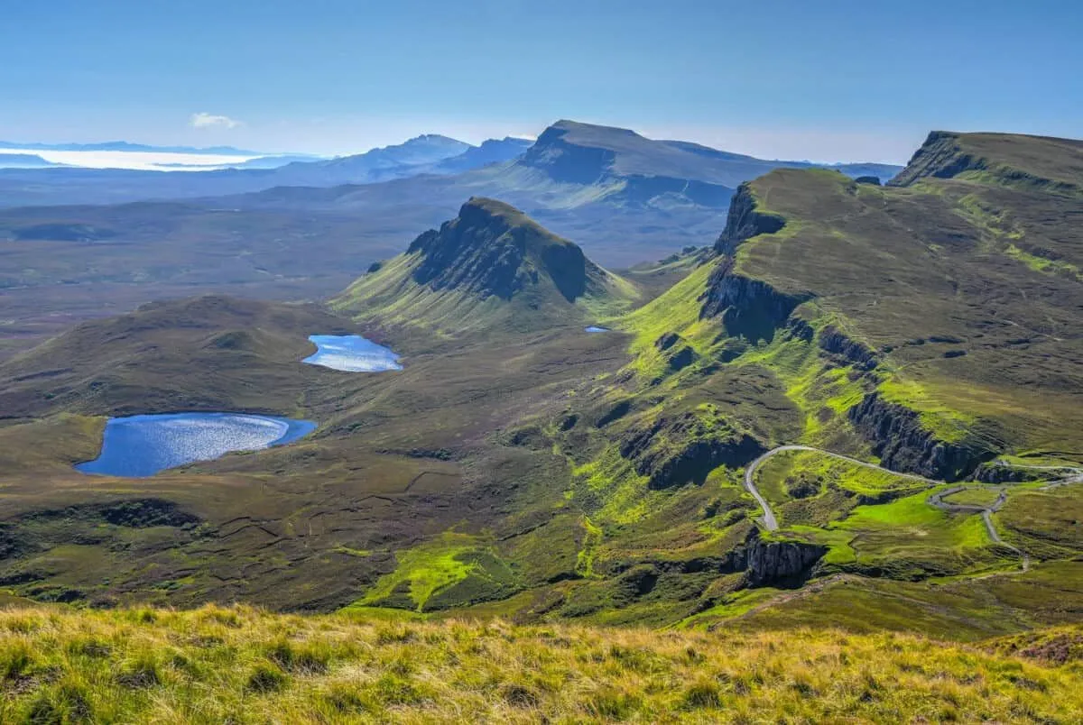 A high-quality photograph of a vast mountainous landscape featuring lush green hills, serene blue lakes, and a winding road under a clear sky. The image captures the natural beauty and grandeur of the outdoors.