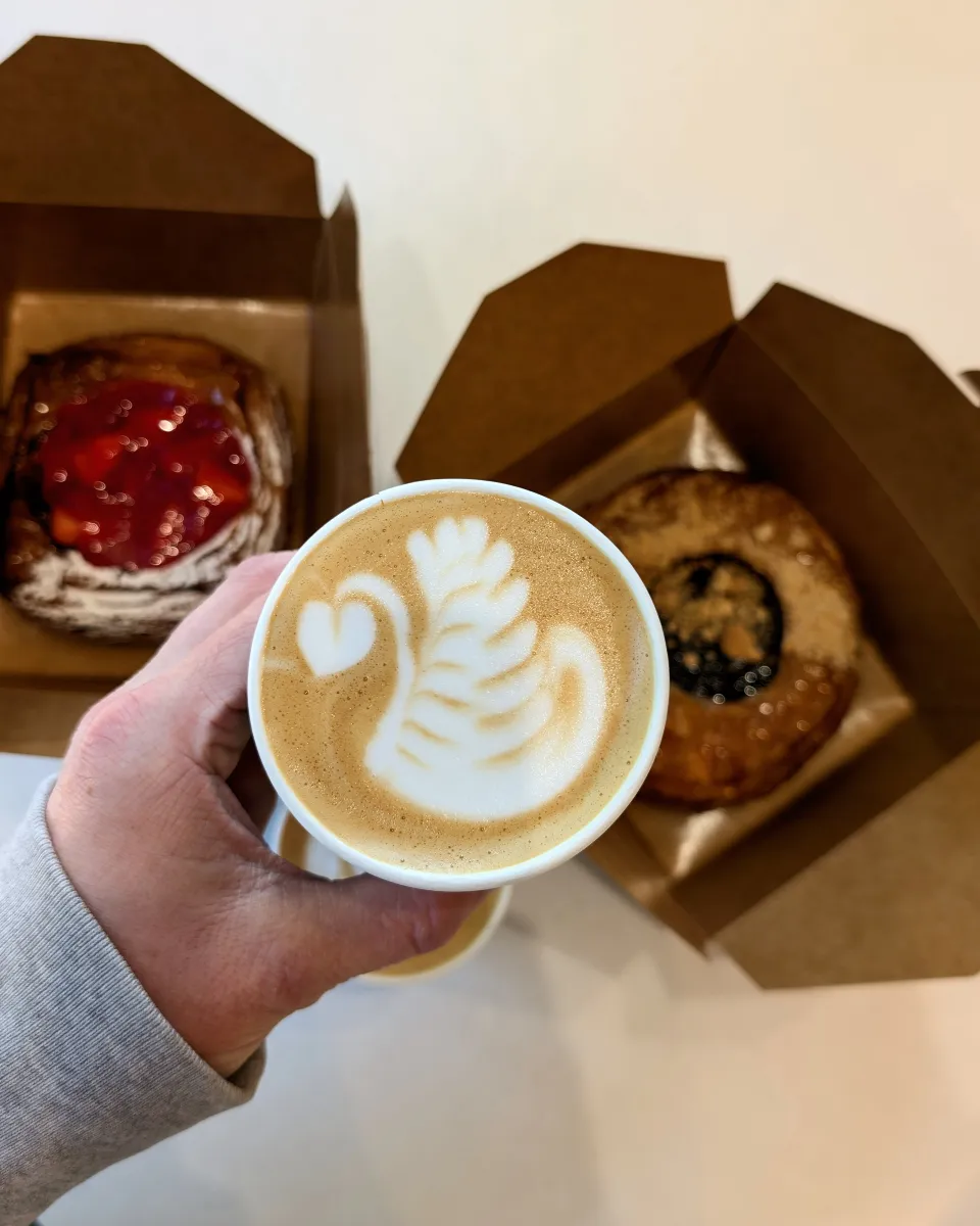 A top-down view of a hand holding a coffee cup with intricate swan latte art, with two pastries in cardboard boxes in the blurred background.