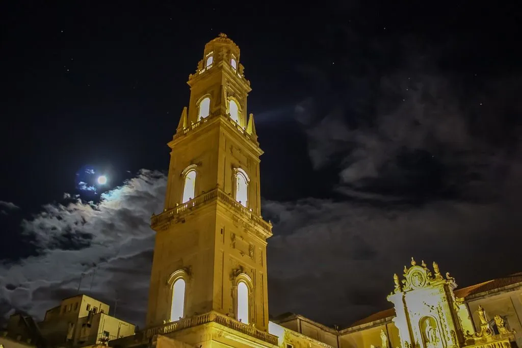 A dramatic night shot of an illuminated historical tower, featuring the moon and clouds in the dark sky.