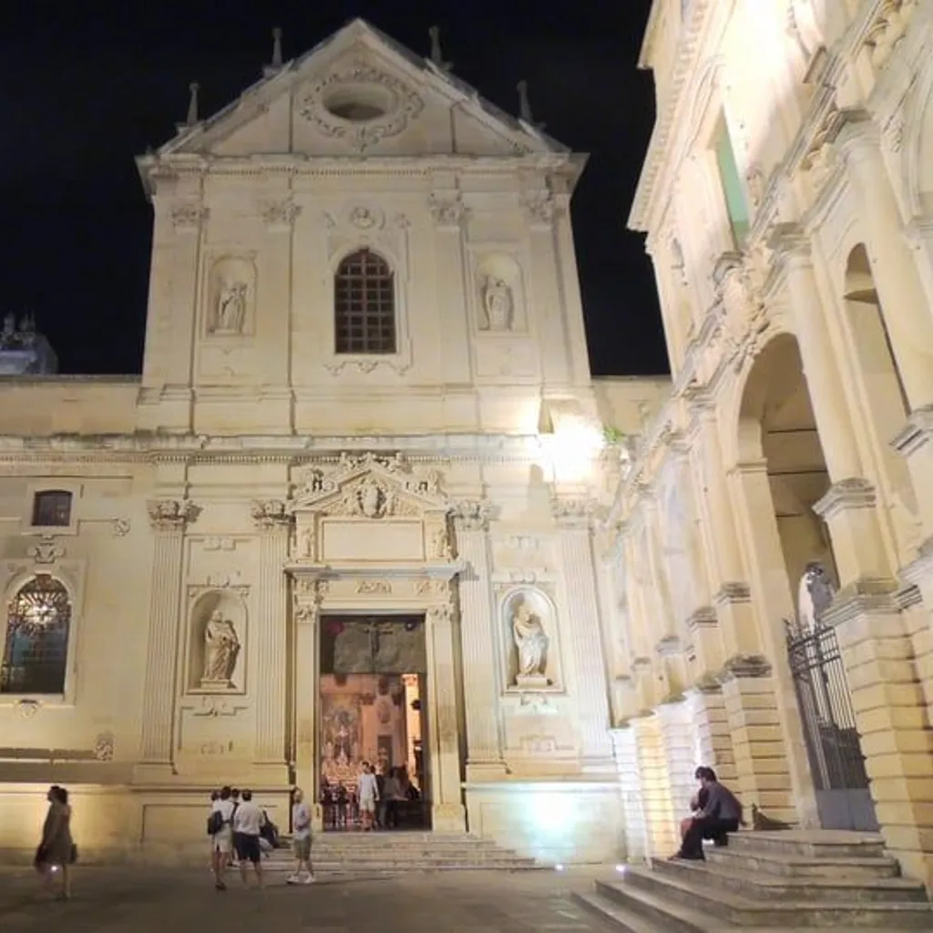 An evening view of a grand, ornately designed Baroque church facade, illuminated by artificial lights against a dark night sky, with a few people gathered in the plaza below.