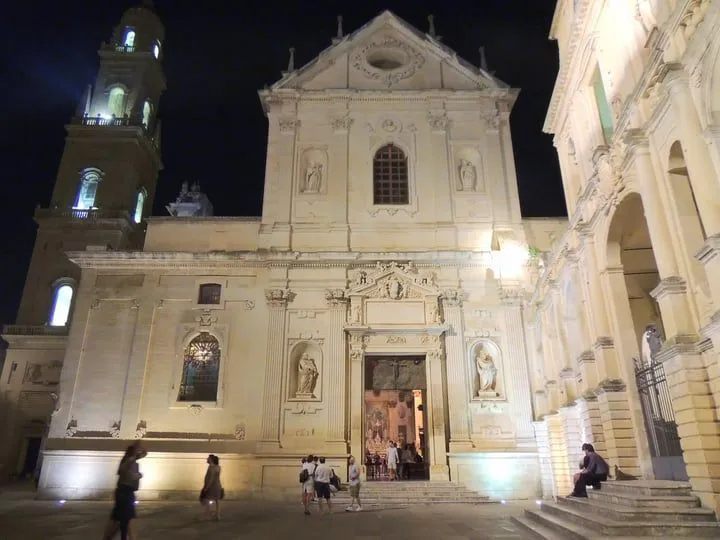 An evening view of a grand, ornately designed Baroque church facade, illuminated by artificial lights against a dark night sky, with a few people gathered in the plaza below.