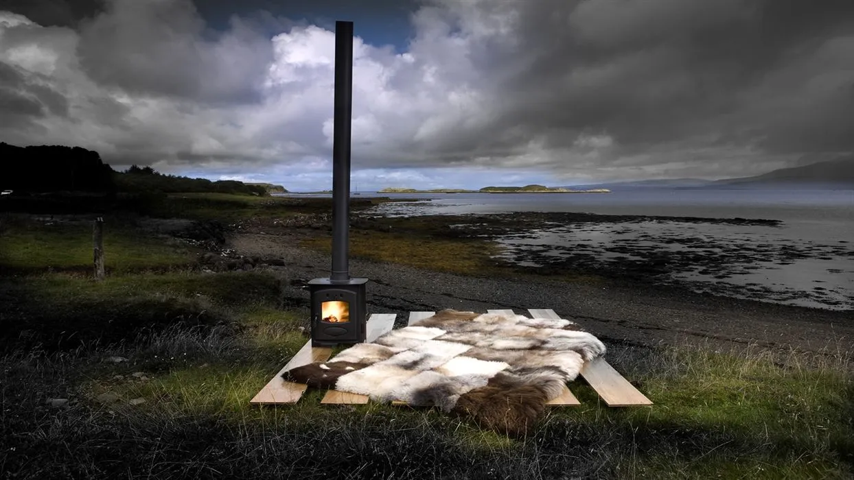 The image features a unique outdoor setup with a wood-burning stove and a fur rug on a wooden base, set against a dramatic coastal landscape under a cloudy sky. The composition effectively frames the subjects within the vast natural environment.