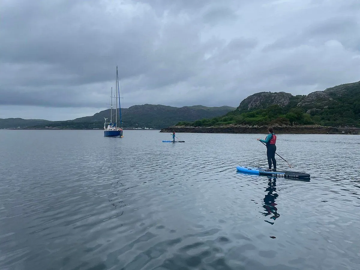 An image featuring two individuals paddleboarding on calm water, with a sailboat anchored in the distance and green hills under an overcast sky.
