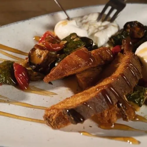 A close-up shot of a food dish featuring toasted bread slices drizzled with sauce, accompanied by cooked vegetables and a creamy white element, possibly cheese or burrata, on a light-colored plate. A fork is visible in the upper right background.