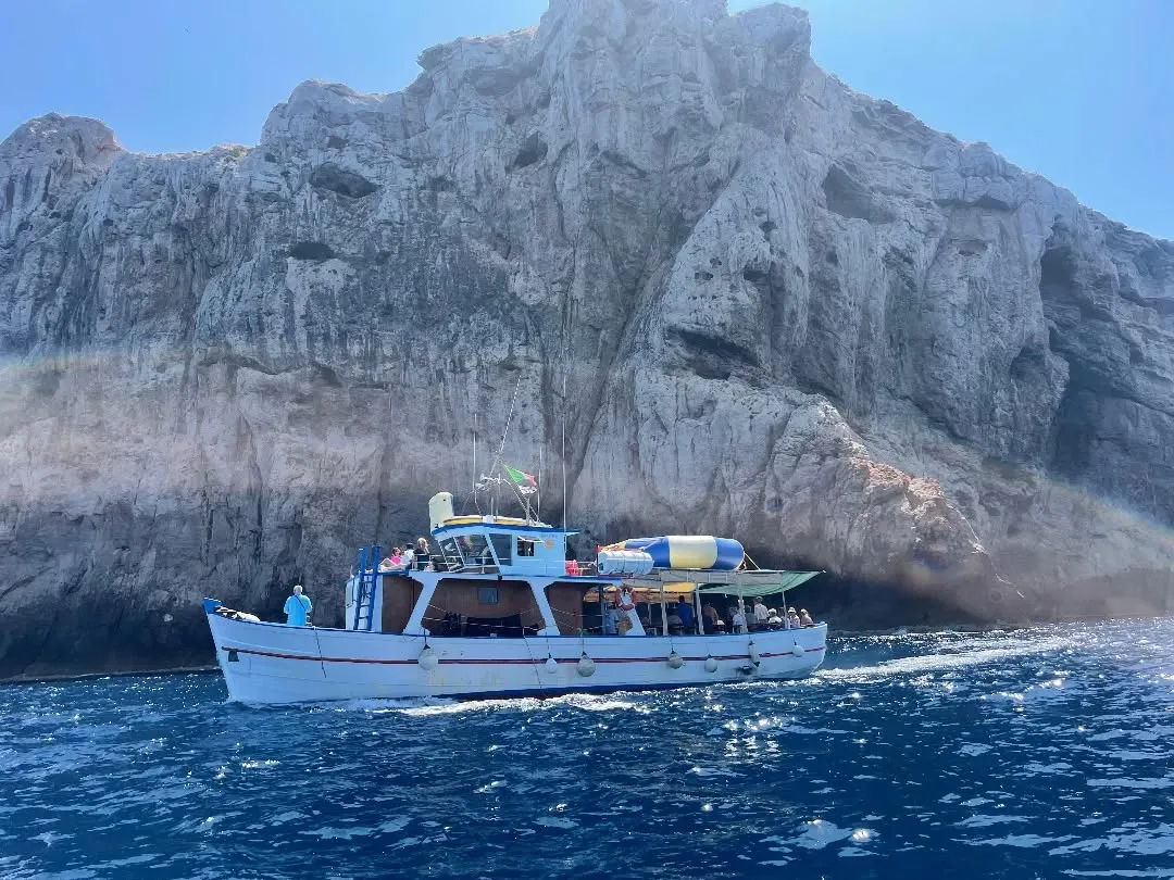 A boat carrying passengers cruises past a large rocky cliff or island on a sunny day, with clear blue water.