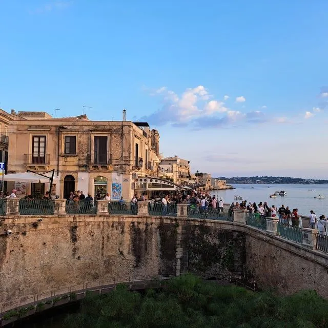 An editorial-style image capturing a vibrant evening scene along a historic waterfront promenade, featuring old European buildings and a crowd of people enjoying the atmosphere.
