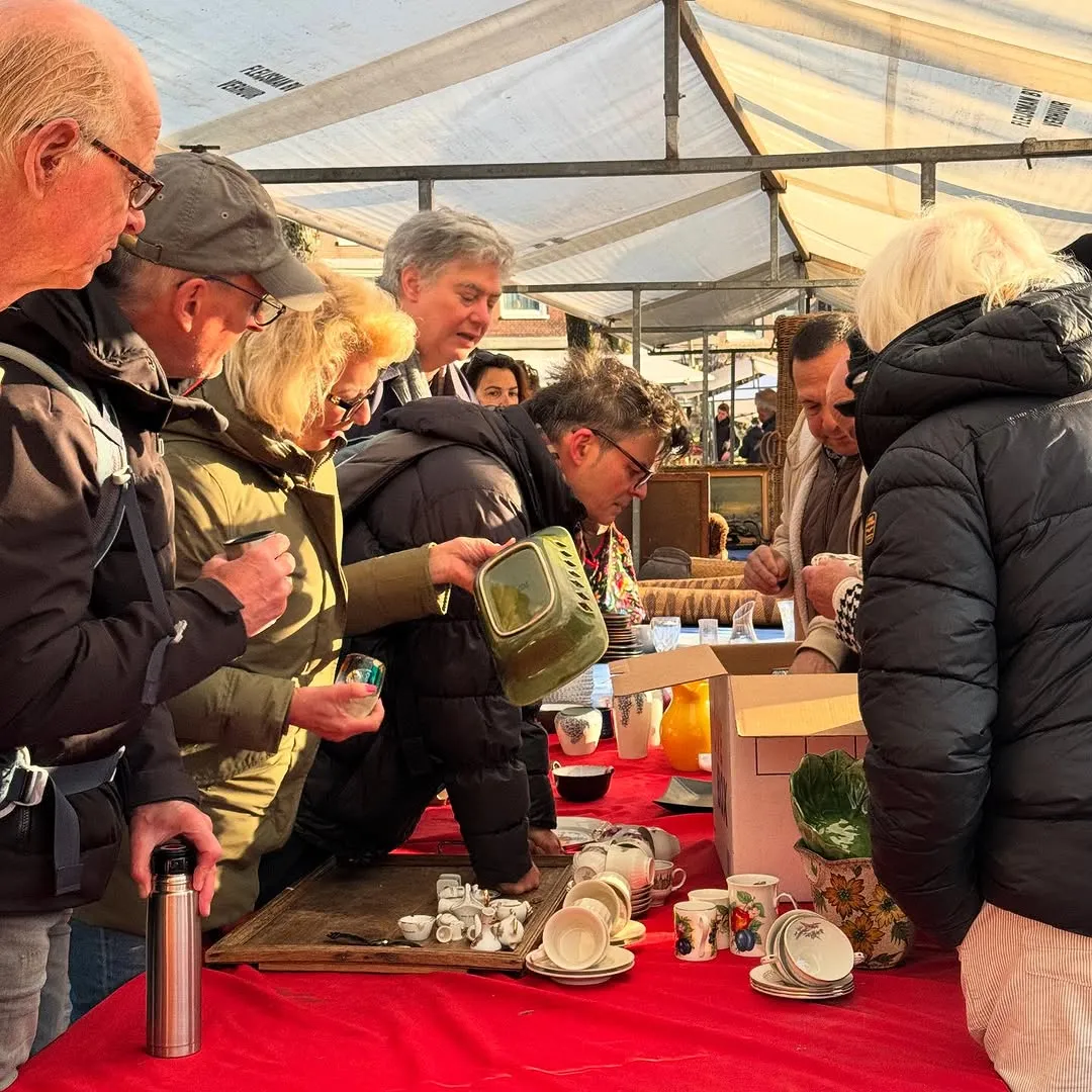 An outdoor market scene showing several people browsing a stall filled with crockery and other items. The image captures the interaction and activity of the shoppers under natural daylight.