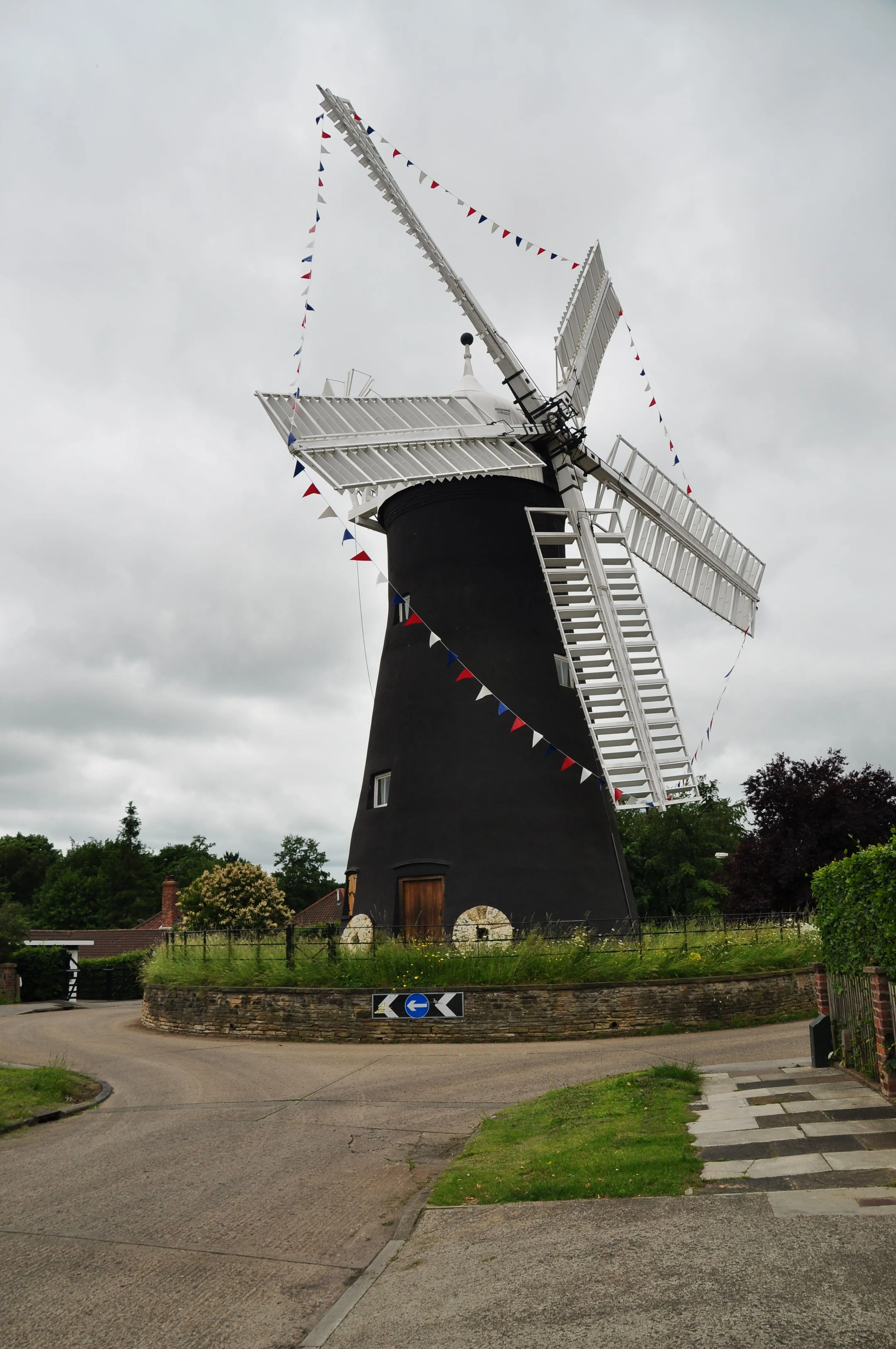 A photograph of a black windmill with white sails adorned with colorful bunting, situated by a road under an overcast sky.
