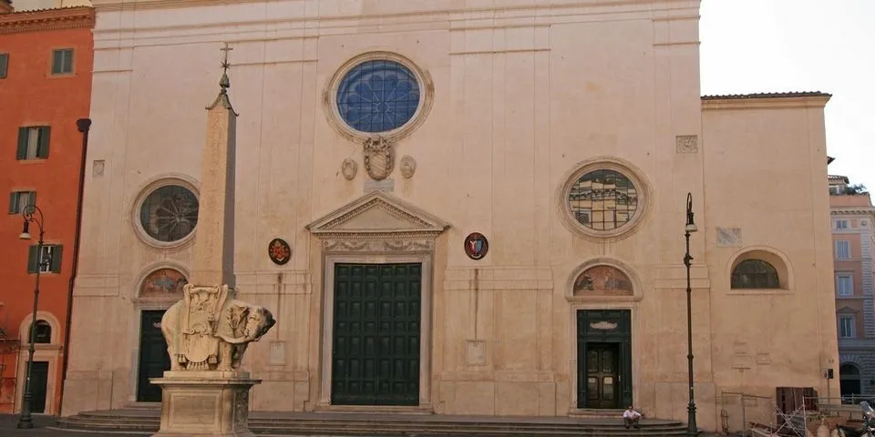 An editorial image showcasing the ornate facade of a historic building, likely a church, with large wooden doors, decorative windows, and carved stone details. A small statue is partially visible on the left, and a person is seated subtly on the steps in the lower right.