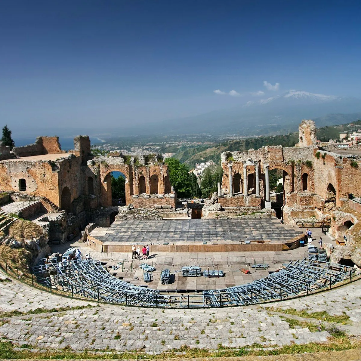 An impressive wide-angle shot of the ancient Greek theater in Taormina, Sicily, with Mount Etna visible in the distant background under a clear blue sky. The image captures the grandeur of the ruins, including the stage area with temporary modern event setups.
