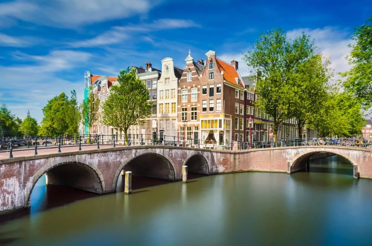An iconic view of a canal in Amsterdam, featuring traditional Dutch canal houses and bridges under a clear blue sky. The water exhibits a smooth, long-exposure effect, contributing to the serene atmosphere of the urban landscape.