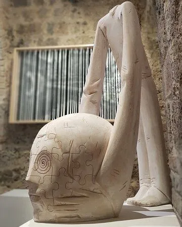The image prominently features a sculpture of a figure with a head resembling a puzzle, positioned against a textured wall and a framed artwork in the background. The composition effectively highlights the unique details of the sculpture.