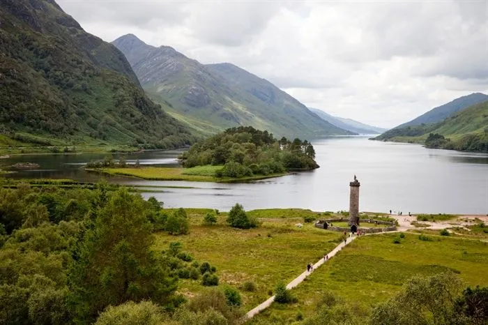 A scenic landscape photograph featuring a loch surrounded by mountains, an island densely covered with trees, and a historic monument with a path leading to it. The sky is overcast, providing even illumination to the scene.