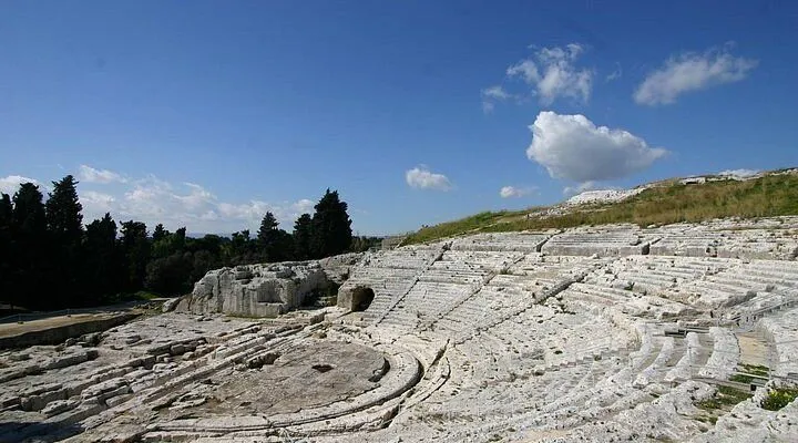 A wide shot of an ancient, open-air theater carved from light-colored stone, featuring tiered seating and a central stage area, set against a vibrant blue sky with scattered white clouds. The historical site is surrounded by green hills and dark trees on the left.