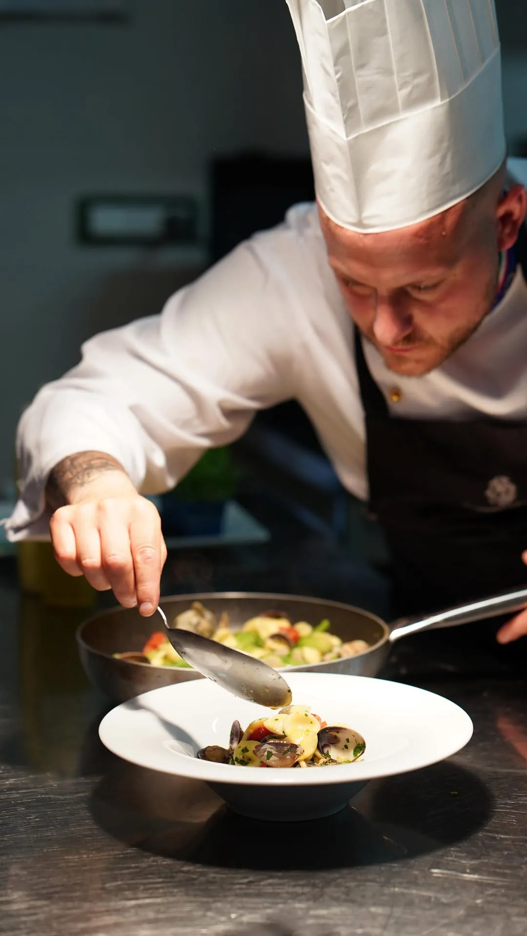 This image showcases a chef meticulously plating a dish, capturing the essence of culinary preparation. The focus is sharp on the chef's hands, the spoon, and the food being served, drawing the viewer's eye to the intricate details of the process. The lighting effectively highlights the main subject, and the colors are natural and accurate. The overall photo-realism is high, making it a compelling visual of a professional kitchen environment.
