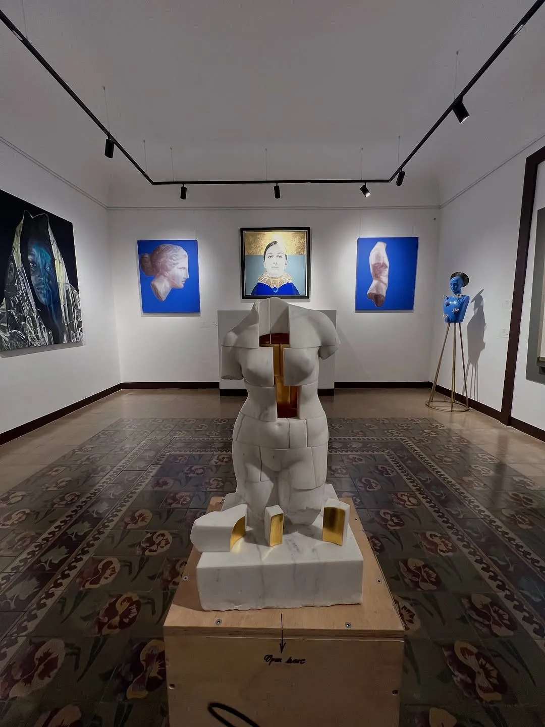 The image is very sharp with excellent focus on both the foreground sculpture and the background paintings, contributing to its high photo-realism. It is well-lit, effectively illuminating the artworks and the space. Colors appear accurate and vibrant. The composition is generally good, showcasing the art effectively, though it feels slightly wide-angle, and there are minor reflections on the polished floor.