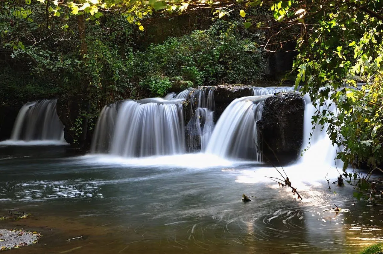 An editorial-style photograph of a multi-tiered waterfall in a lush green environment. The long exposure technique renders the flowing water with a smooth, silky effect, contrasting with the sharp details of the surrounding rocks and foliage. The lighting is soft and natural, contributing to a serene and realistic natural scene.