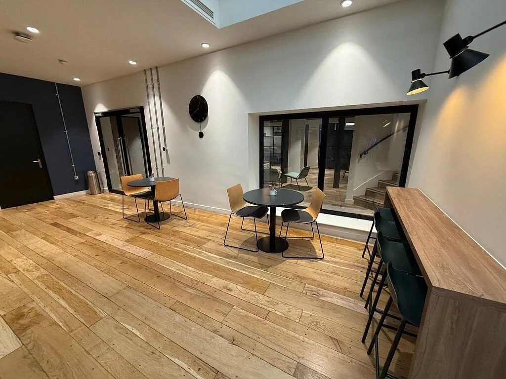 The image features a contemporary interior space with wooden floors, small round tables with chairs, and a bar counter. The lighting is artificial, creating bright spots on the walls, and the space appears clean and well-maintained. A large window or glass partition reveals a staircase in the background.