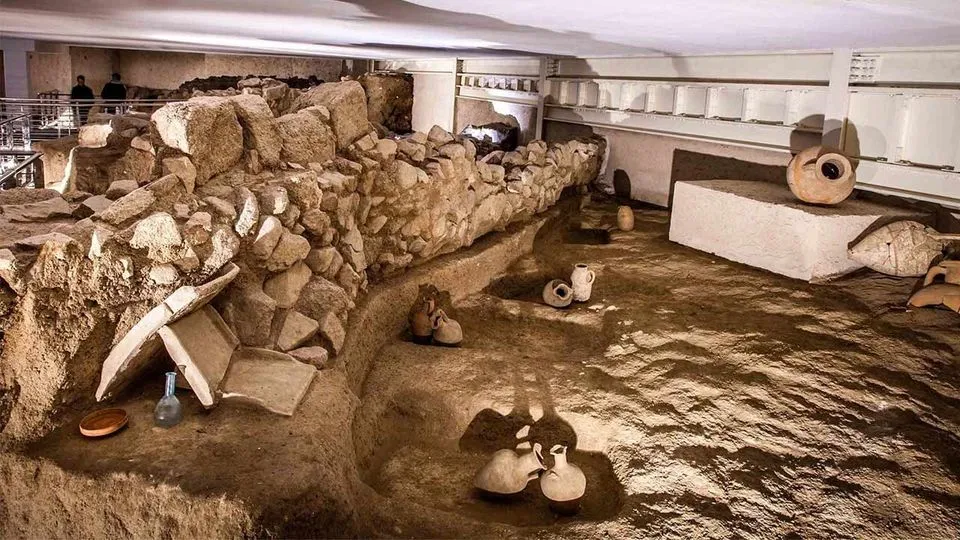 The image depicts an archaeological excavation site, featuring excavated stone walls and several ancient pottery vessels. The composition is strong, offering a clear view of the site with good depth, and the key elements are in sharp focus. The image appears photo-realistic and the earthy colors are accurate to the subject matter. The lighting is generally adequate, though a bit flat, which slightly reduces the overall dynamic range and mood.