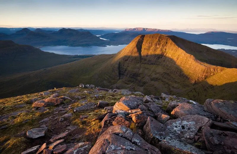 An elevated view of a vast mountainous landscape bathed in golden hour light, featuring rugged foreground rocks, a prominent mountain peak, and a body of water winding through the valley in the distance.