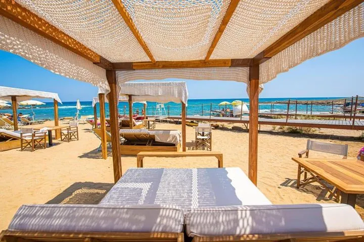 The image is well-composed, using the cabana structure to frame the inviting beach scene and ocean beyond. It is well-lit by natural sunlight, resulting in vibrant and accurate colors. The overall sharpness is excellent, contributing to its high photo-realism, and all key elements are clearly in focus. The strong shadows from the overhead netting add texture and depth to the foreground.