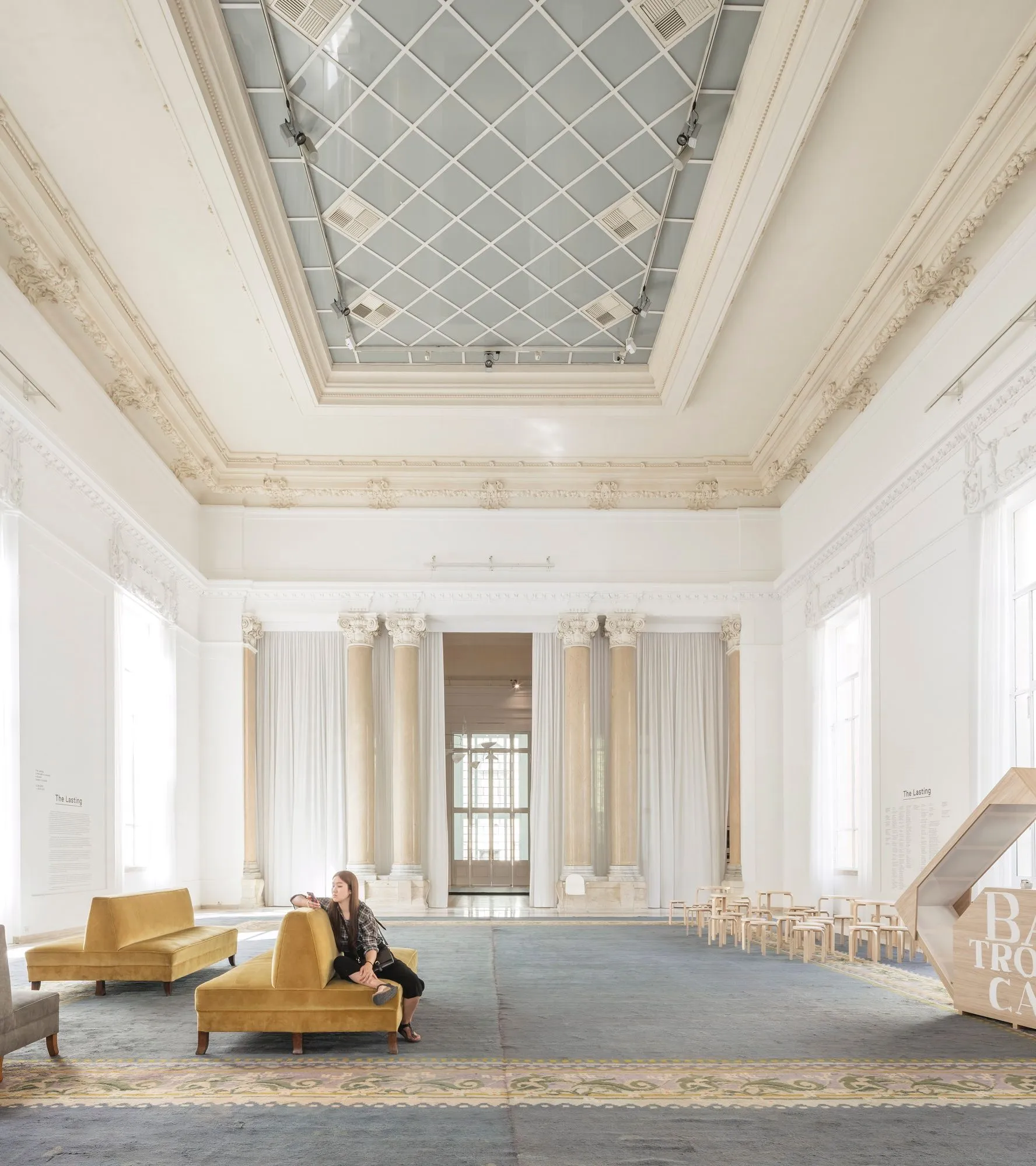 An editorial-style image capturing a grand, well-lit interior space. The room features high ceilings with ornate molding, large windows, and classical columns, suggesting a historical building or museum. The scene includes modern yellow couches and a person seated on one, along with a sculptural wooden element and small chairs, possibly indicating an exhibition or event. The overall composition is clean and visually appealing.