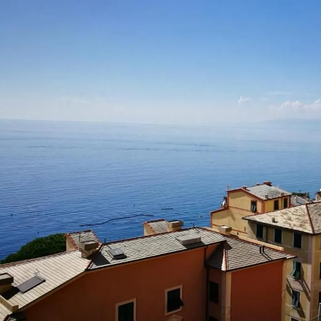 An elevated view of a coastal town with colorful buildings overlooking a clear blue sea under a bright sky. A white decorative railing is visible on the right, framing the picturesque scene with distant mountains along the coast.