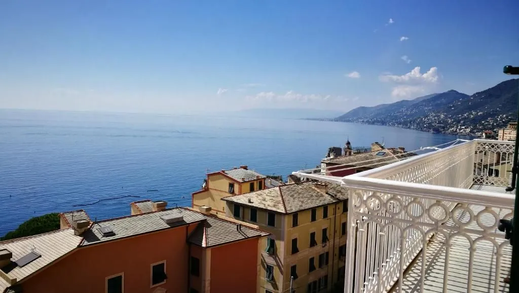 An elevated view of a coastal town with colorful buildings overlooking a clear blue sea under a bright sky. A white decorative railing is visible on the right, framing the picturesque scene with distant mountains along the coast.