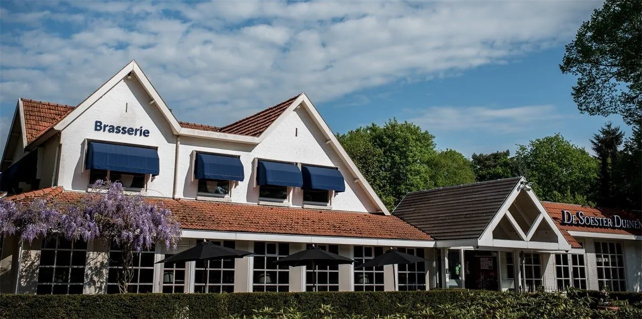 An exterior view of a white building featuring a red-tiled roof, blue awnings over the upper windows, and dark umbrellas positioned in front of the lower windows, set against a bright blue sky with clouds and lush green trees.