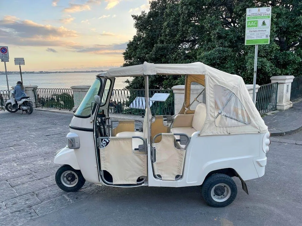 An editorial-style image featuring a vintage-style white three-wheeled vehicle parked by a waterfront. The vehicle is the clear subject, well-lit by natural light, and sharply in focus. The background shows a calm body of water and trees, with a pleasant, soft sky.