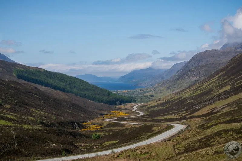 A picturesque landscape featuring a winding road through a valley, leading to a loch and mountains in the distance under a blue sky. The composition effectively draws the eye into the scene, and the natural lighting is well-balanced.