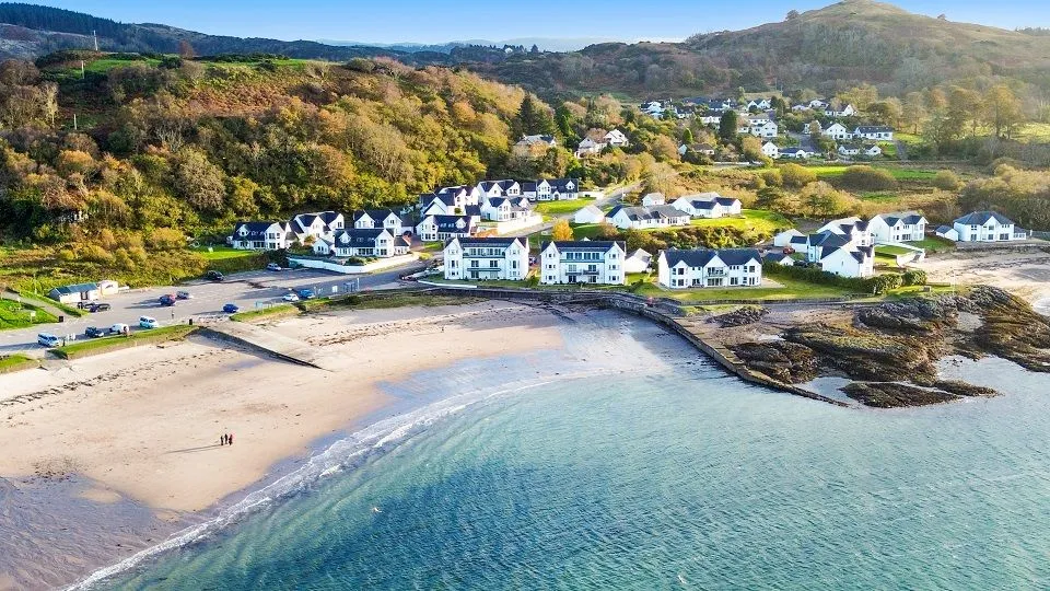 The image is a high-quality, well-composed aerial view of a charming coastal village nestled against hills, featuring a beautiful sandy beach and clear turquoise waters. It benefits from excellent natural lighting, sharp details, accurate colors, and strong photo-realism.