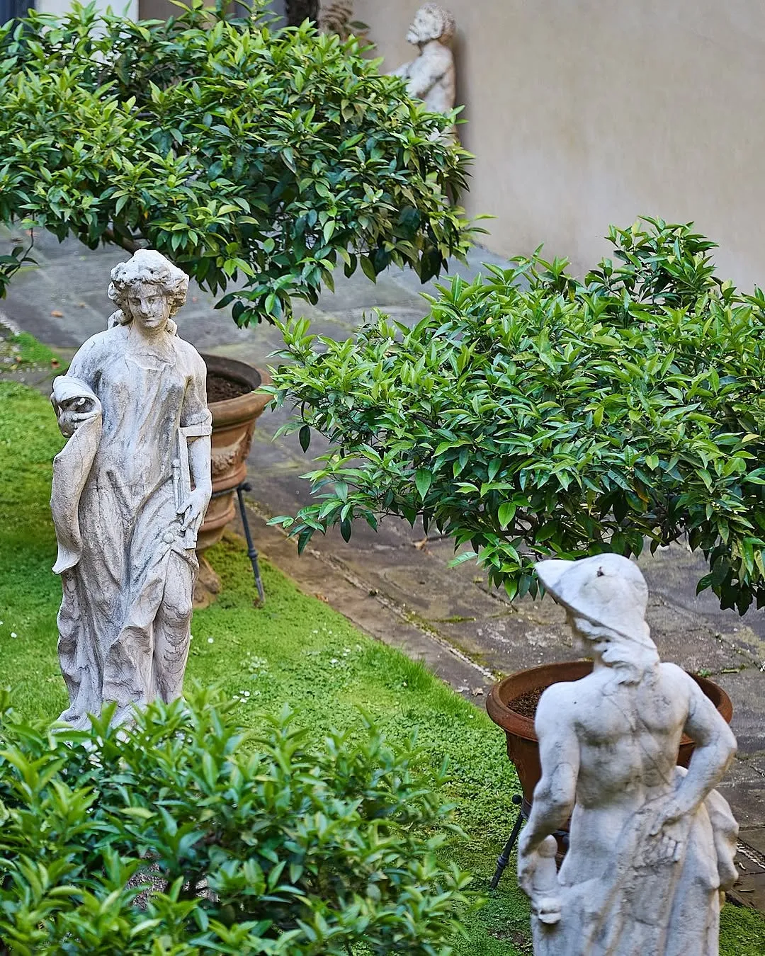 A serene outdoor scene featuring two classical stone statues amidst lush green foliage in a garden setting. A female figure in the foreground stands prominently, while a male figure is partially visible in the mid-ground, both surrounded by potted plants and garden paths.