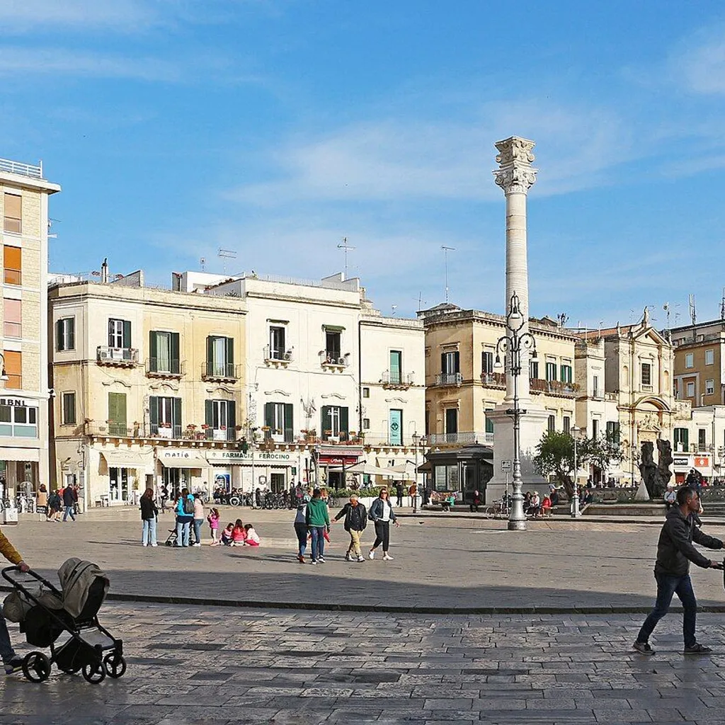 An editorial style image showcasing a bustling city square in Italy, featuring historic buildings, a prominent column, and people walking about under a clear blue sky.