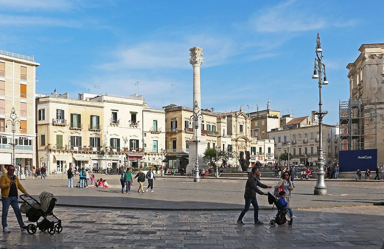 An editorial style image showcasing a bustling city square in Italy, featuring historic buildings, a prominent column, and people walking about under a clear blue sky.