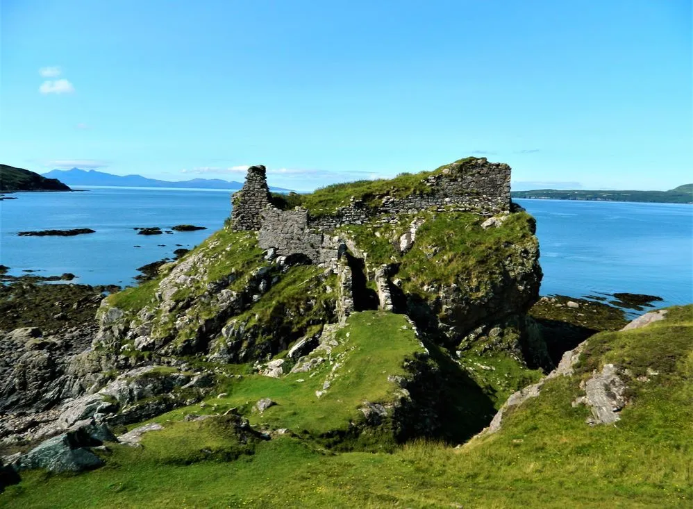 An editorial quality image depicting ancient castle ruins situated on a rocky, grassy outcrop overlooking a vast expanse of blue ocean under a clear sky. The image captures the rugged beauty of the coastal landscape with historical elements.
