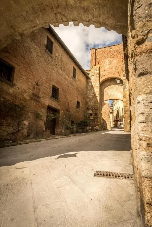 A low-angle view of a narrow, sunlit street in an old European town, flanked by tall brick and stone buildings. An ancient archway in the midground opens to reveal more historic architecture down the winding street.