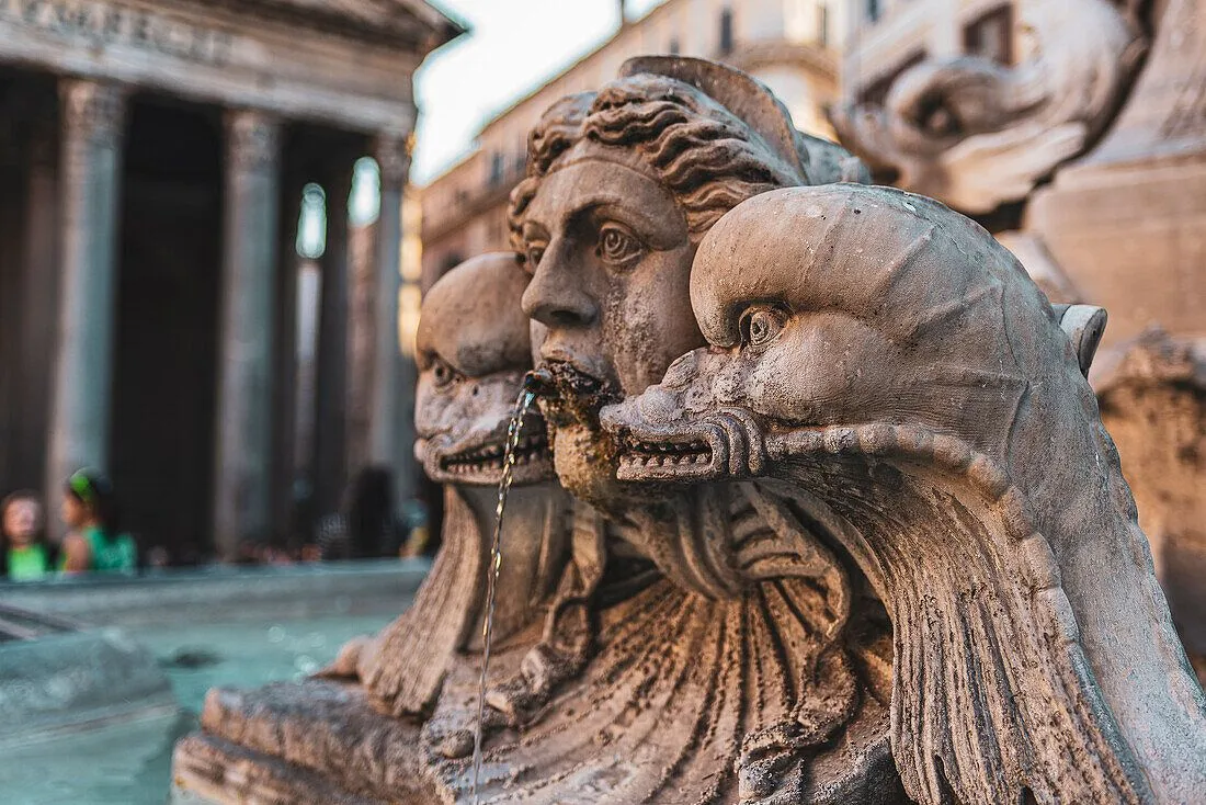 A close-up photograph of a detailed stone fountain sculpture, depicting a mythological figure's face with water flowing from its mouth, flanked by stylized sea creatures. The background shows blurred architectural elements, possibly a historical building.