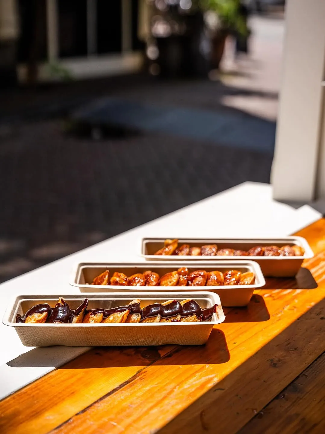 The image is well-composed with a pleasing diagonal arrangement of the food items. The natural sunlight creates good contrast and highlights the texture of the churros and sauces, making them look appealing. The foreground churros are sharp and in excellent focus, contributing to the photo-realistic quality. The colors are vibrant and accurate. While the lighting is generally good, some areas of the background and the churros further back are slightly less illuminated and sharp due to depth of field, but this is an aesthetic choice.