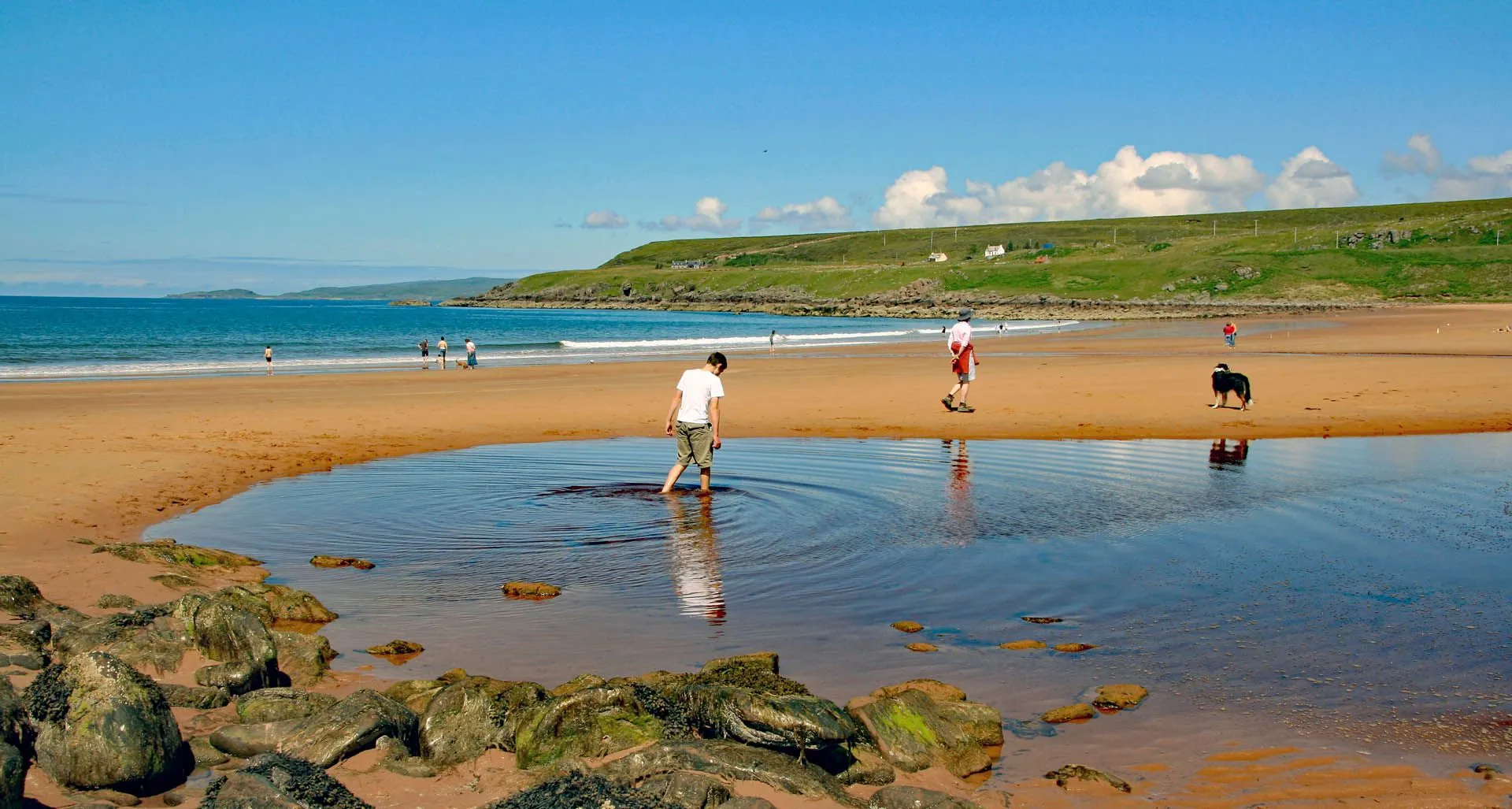 A vibrant and well-composed image capturing a sunny day at the beach. The scene features a person wading in a tidal pool in the foreground, with reflections visible in the water. Beyond the pool, the golden sandy beach stretches towards the blue sea and distant green hills under a bright blue sky with scattered clouds. Other people are visible further down the beach.