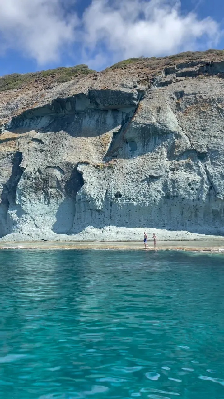 A striking landscape photograph featuring a massive, textured light-colored cliff face rising from clear, turquoise water. Two small human figures are visible walking on a narrow strip of land at the base of the cliff.
