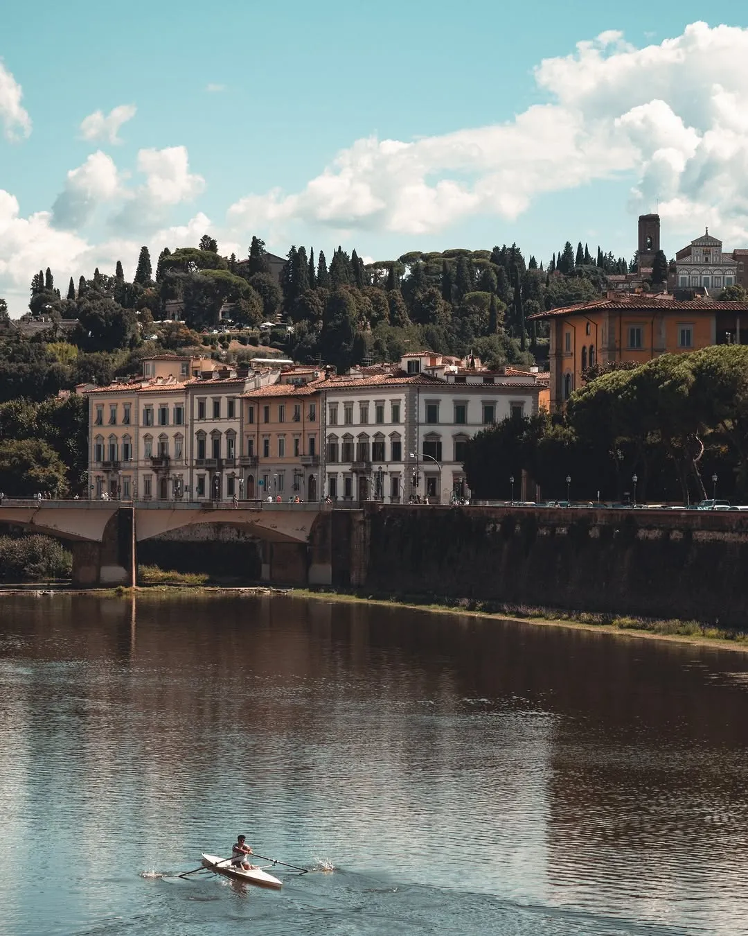 A picturesque view of a historic European city, featuring old buildings along a river, spanned by a classical bridge, under a bright, cloudy sky.