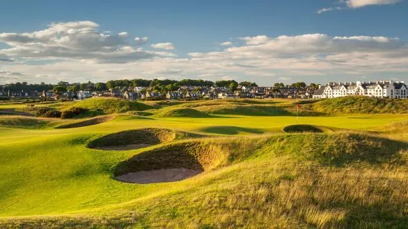 A scenic view of a golf course featuring prominent sand bunkers and a lush green fairway, set against a backdrop of distant residential buildings and a blue sky with scattered clouds. The scene conveys an open, recreational atmosphere.