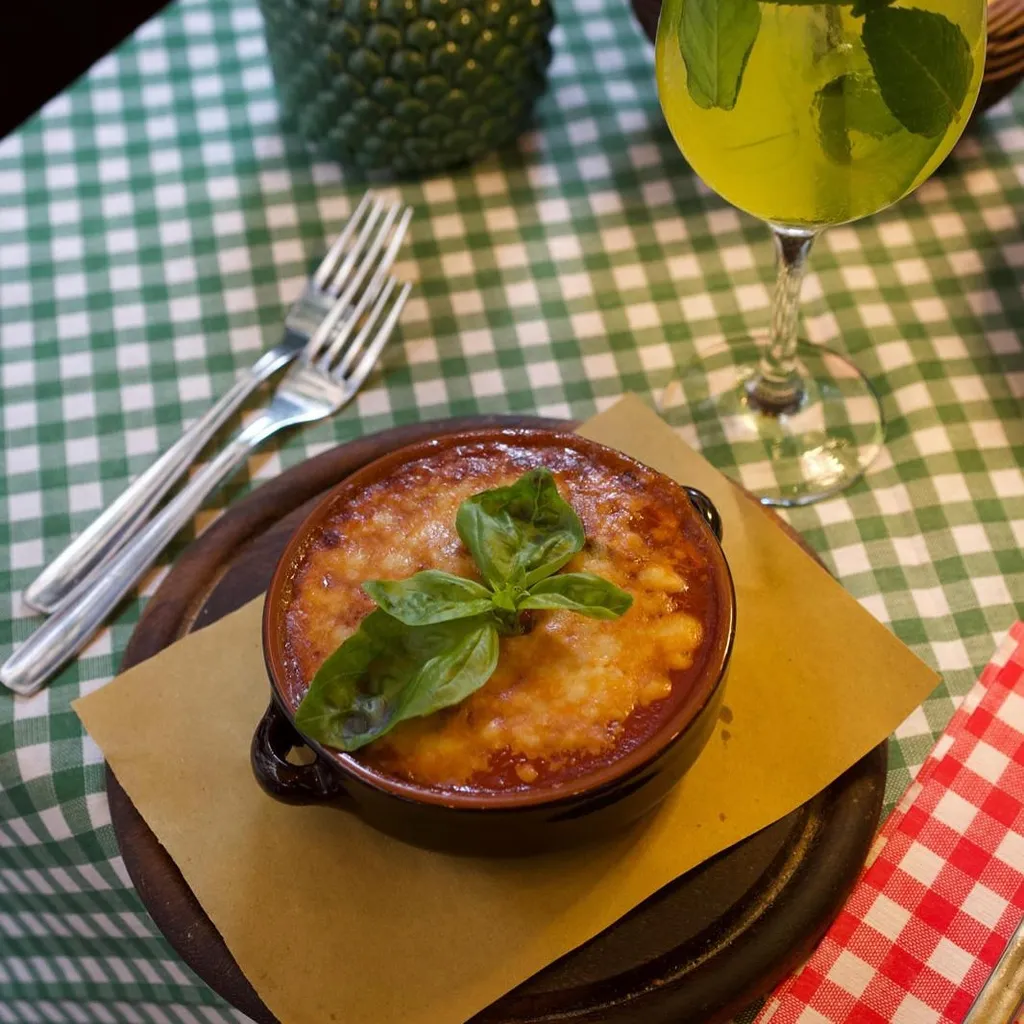The image features a well-composed shot of a baked dish, likely pasta or a casserole, garnished with basil. The main subject is sharp and well-lit, making the food look appetizing and photo-realistic. Colors are accurate and vibrant, especially the green basil and the checkered tablecloth. The key elements, particularly the food, are clearly in focus. While the composition is strong, the background elements like the napkin holder and basket, along with the checkered tablecloth, contribute to a slightly busy environment, though not to the point of being explicitly 'cluttered' or distracting from the main subject.
