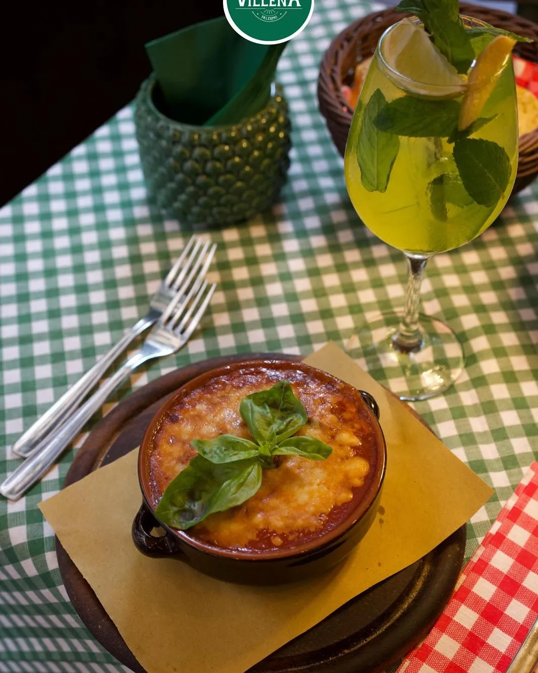The image features a well-composed shot of a baked dish, likely pasta or a casserole, garnished with basil. The main subject is sharp and well-lit, making the food look appetizing and photo-realistic. Colors are accurate and vibrant, especially the green basil and the checkered tablecloth. The key elements, particularly the food, are clearly in focus. While the composition is strong, the background elements like the napkin holder and basket, along with the checkered tablecloth, contribute to a slightly busy environment, though not to the point of being explicitly 'cluttered' or distracting from the main subject.