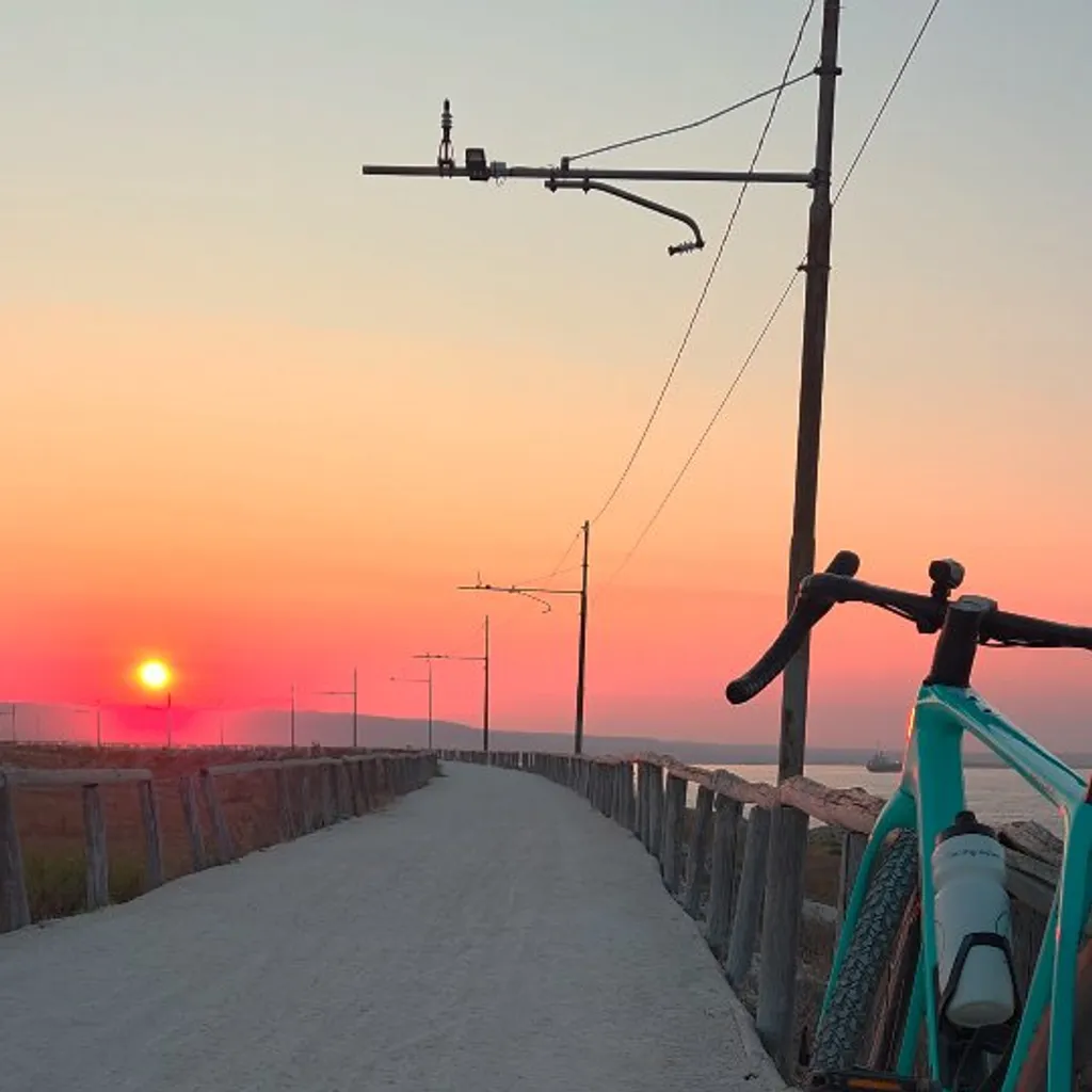 The image features a bicycle parked on a pathway during sunset, creating a serene and adventurous mood. The composition effectively uses leading lines from the path and fence to draw the eye towards the vibrant sunset in the background. The bike in the foreground provides good depth and context. The scene is highly photo-realistic, capturing the natural beauty of the moment.