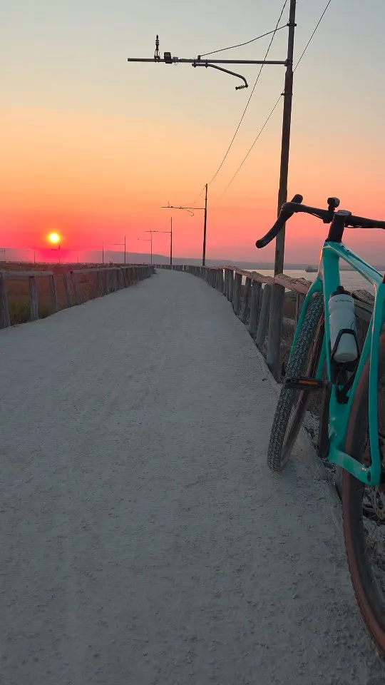 The image features a bicycle parked on a pathway during sunset, creating a serene and adventurous mood. The composition effectively uses leading lines from the path and fence to draw the eye towards the vibrant sunset in the background. The bike in the foreground provides good depth and context. The scene is highly photo-realistic, capturing the natural beauty of the moment.