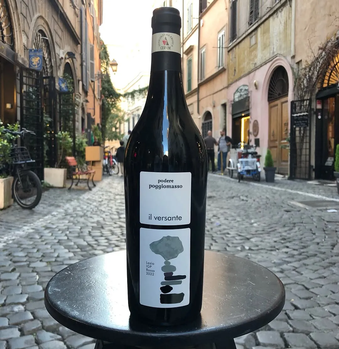A close-up shot of a dark wine bottle positioned on a small dark table on a cobblestone street, with a blurred urban background featuring historic buildings and faint human figures.
